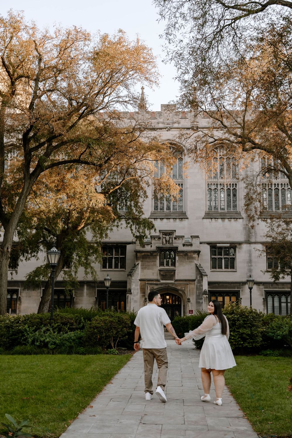 A couple strolls hand in hand along a stone pathway beside a Gothic university building in autumn.