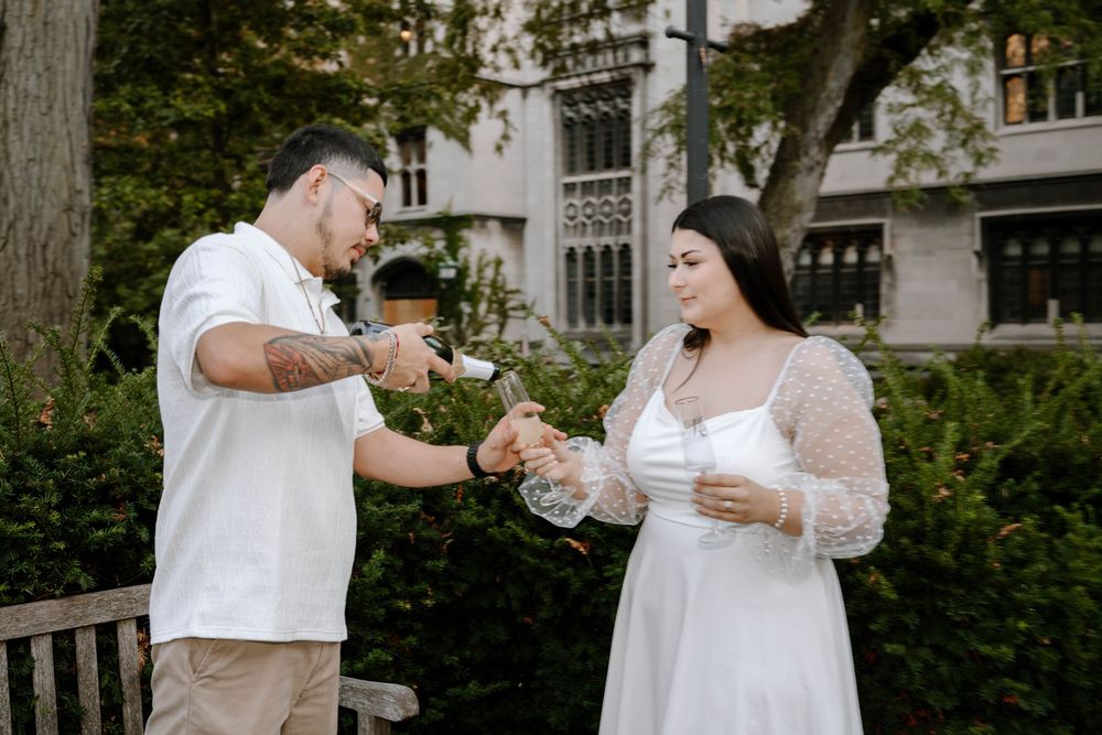 A couple shares drinks together on a bench in a lush garden setting near a historic building.