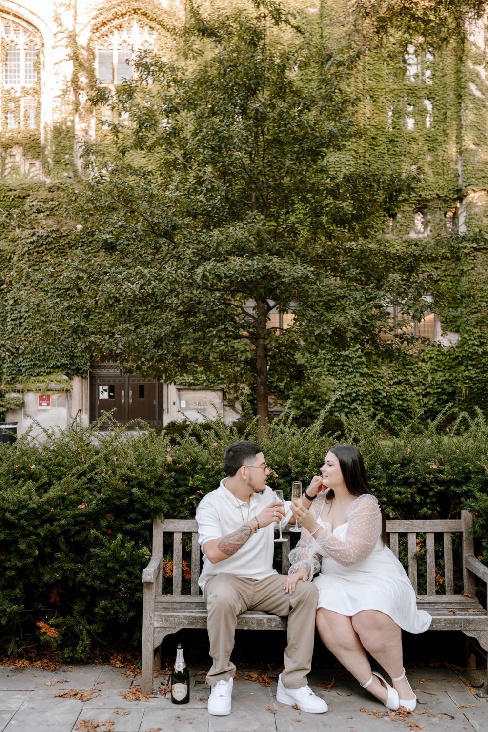 Two people share champagne while sitting on a wooden bench in a garden setting.