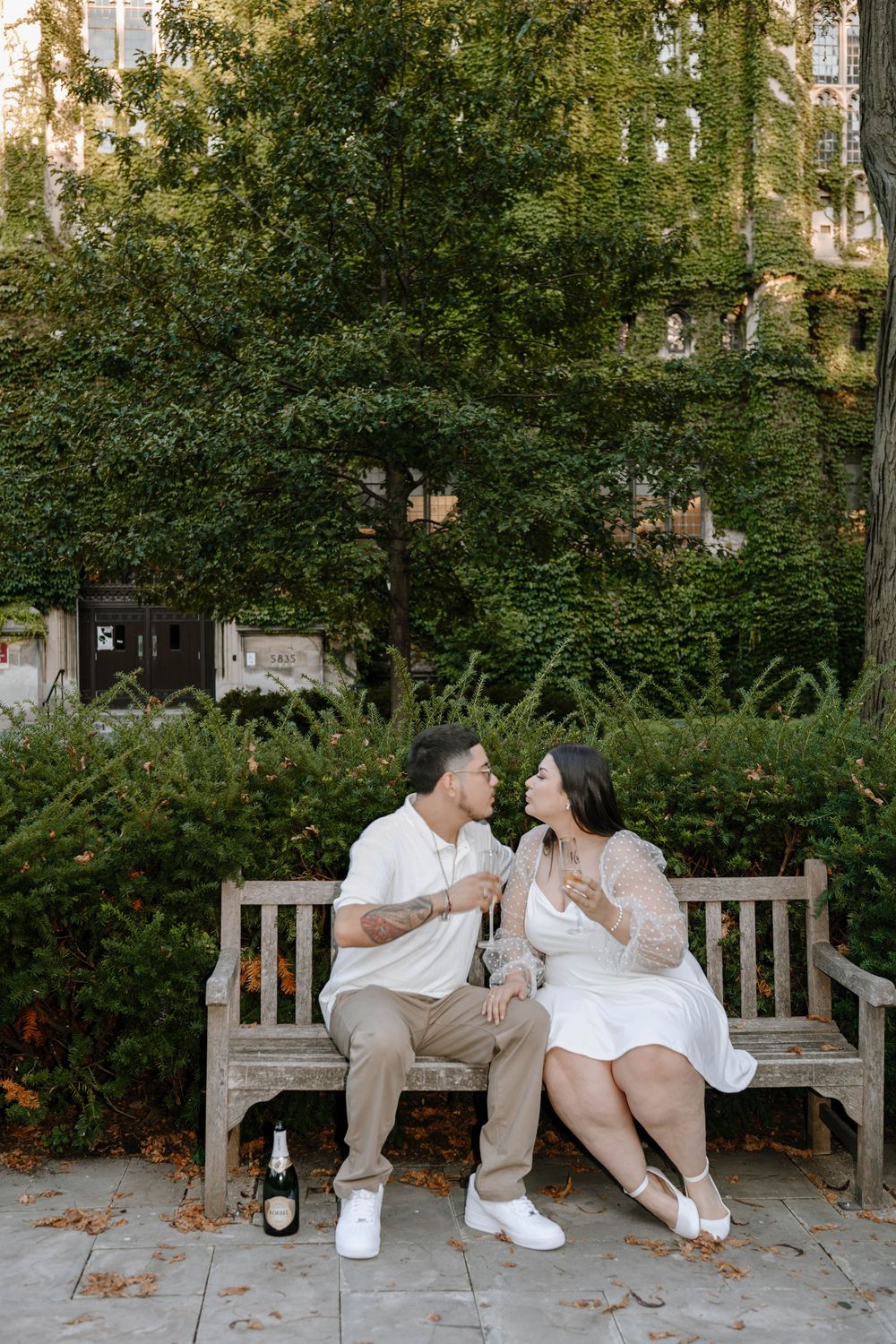 A couple shares champagne while sitting on a wooden bench in a garden setting.