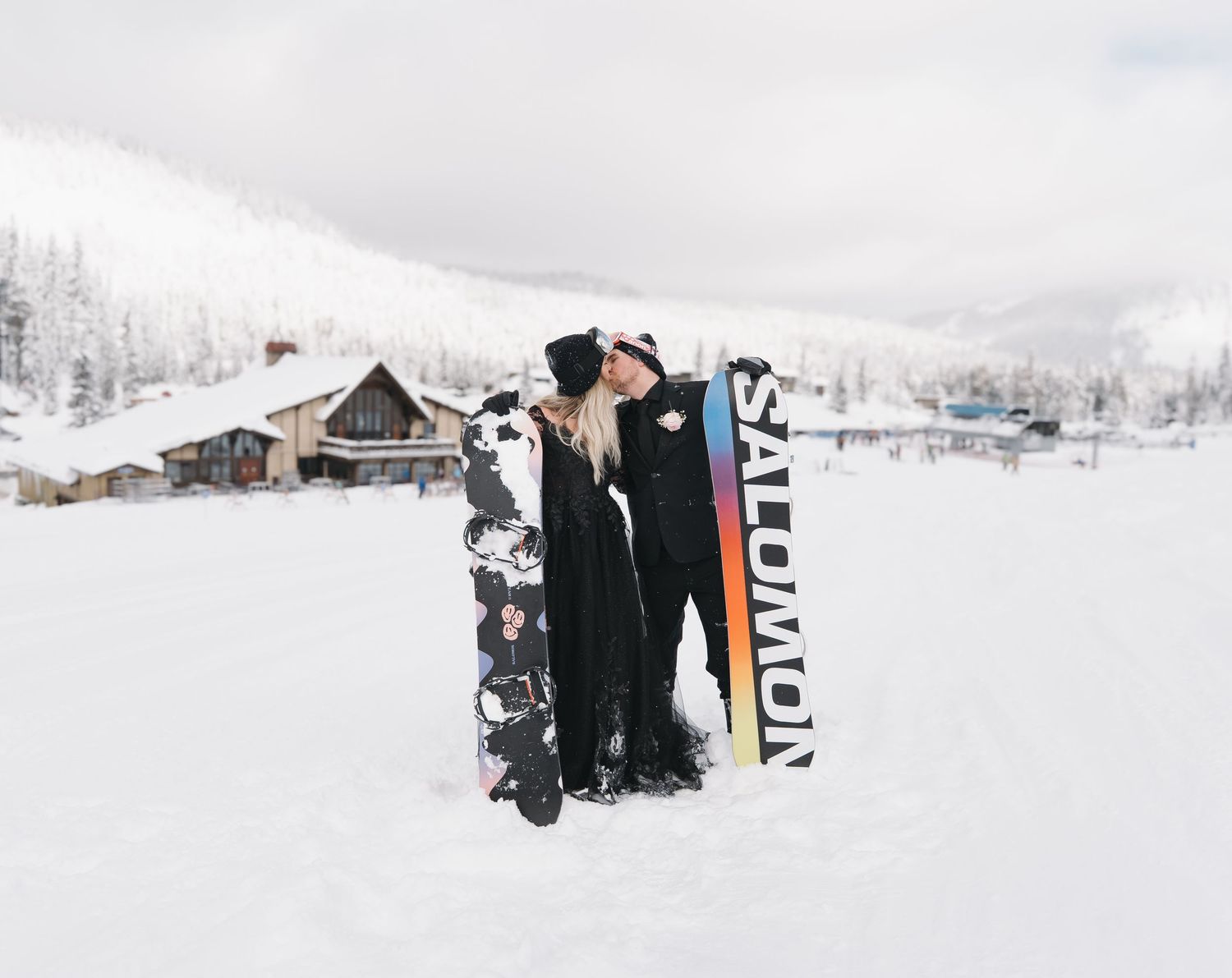 Snowboarders stand together on a snowy slope at a winter resort with snow-covered mountains in the background.