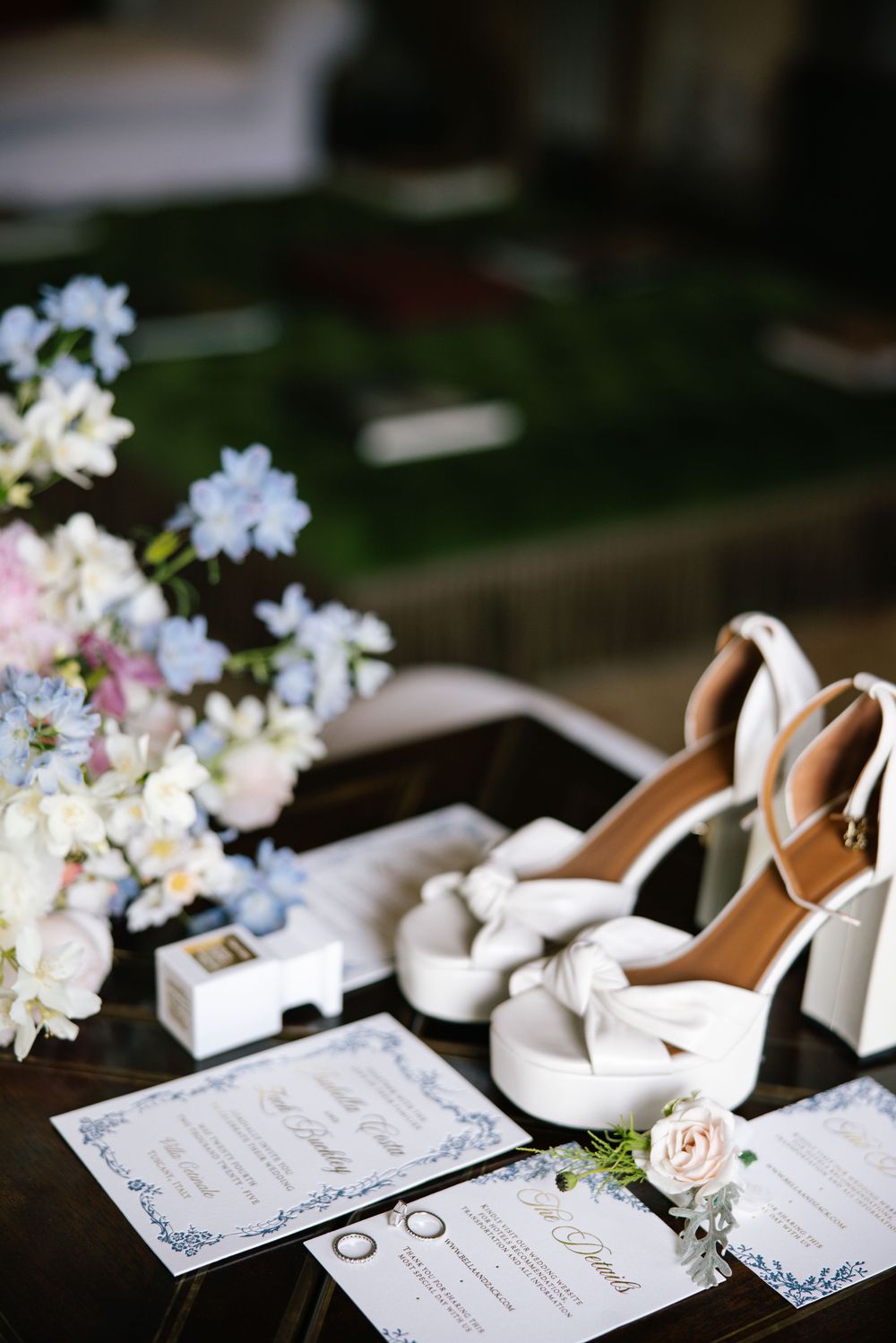 Wedding details including white bridal shoes, delicate flowers, and invitation cards on dark wood surface.