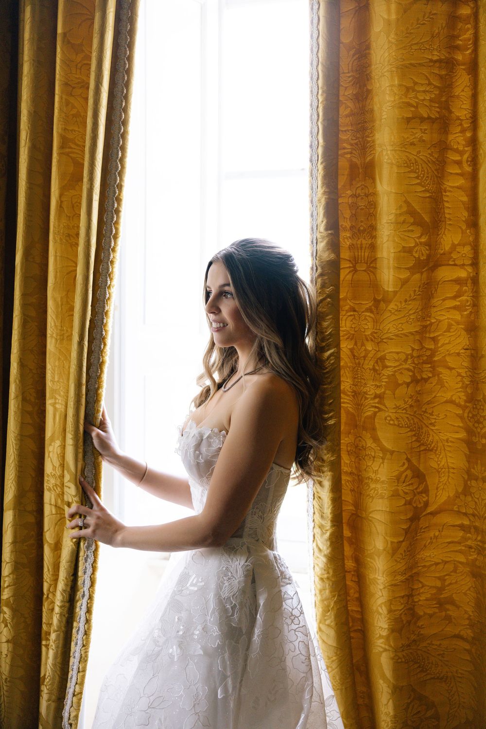 White lace wedding gown silhouetted against golden curtains in natural window light.