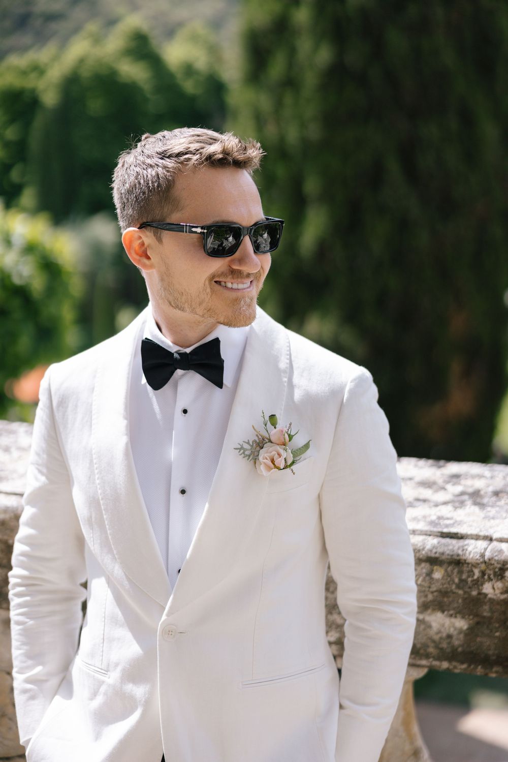 Groom in a crisp white tuxedo and black bowtie smiles while wearing sunglasses outdoors.