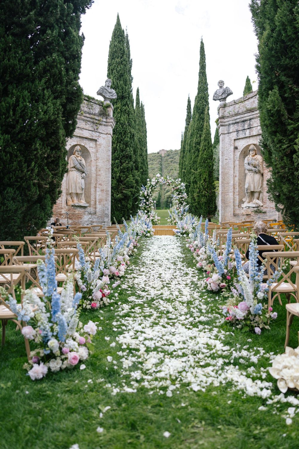 Garden wedding ceremony setup with white flower petals and blue delphinium lining a stone-walled pathway.