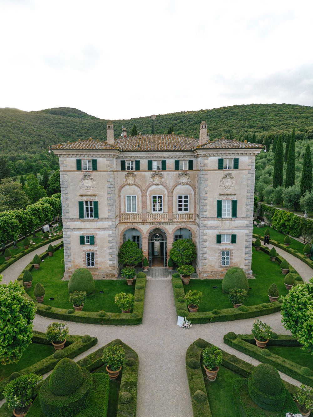 Aerial drone views of a grand Mediterranean villa with manicured gardens and symmetrical pathways in a lush hillside setting.