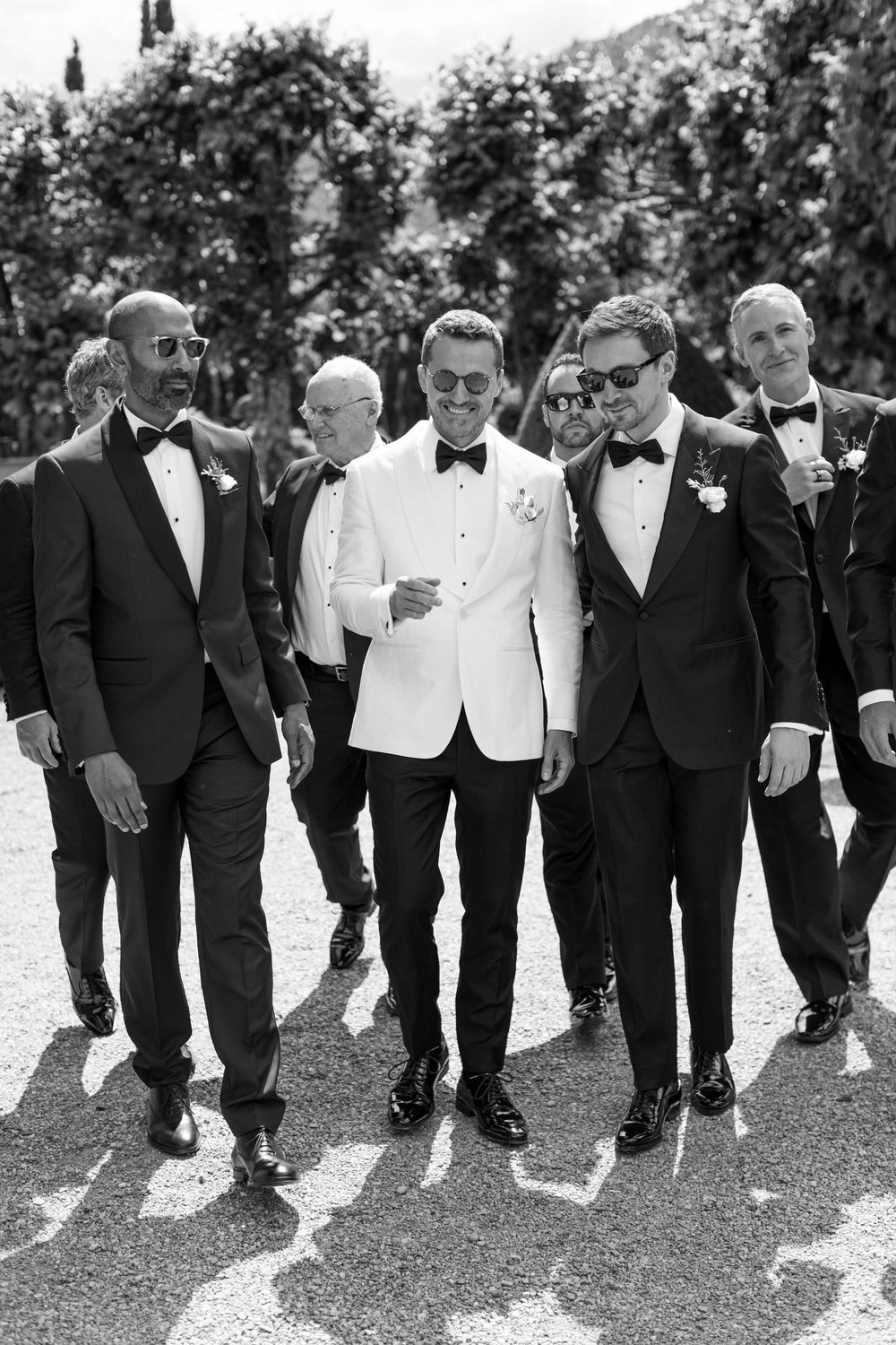 Wedding party in black and white tuxedos walk together in a line looking down at their shoes during outdoor photo session.
