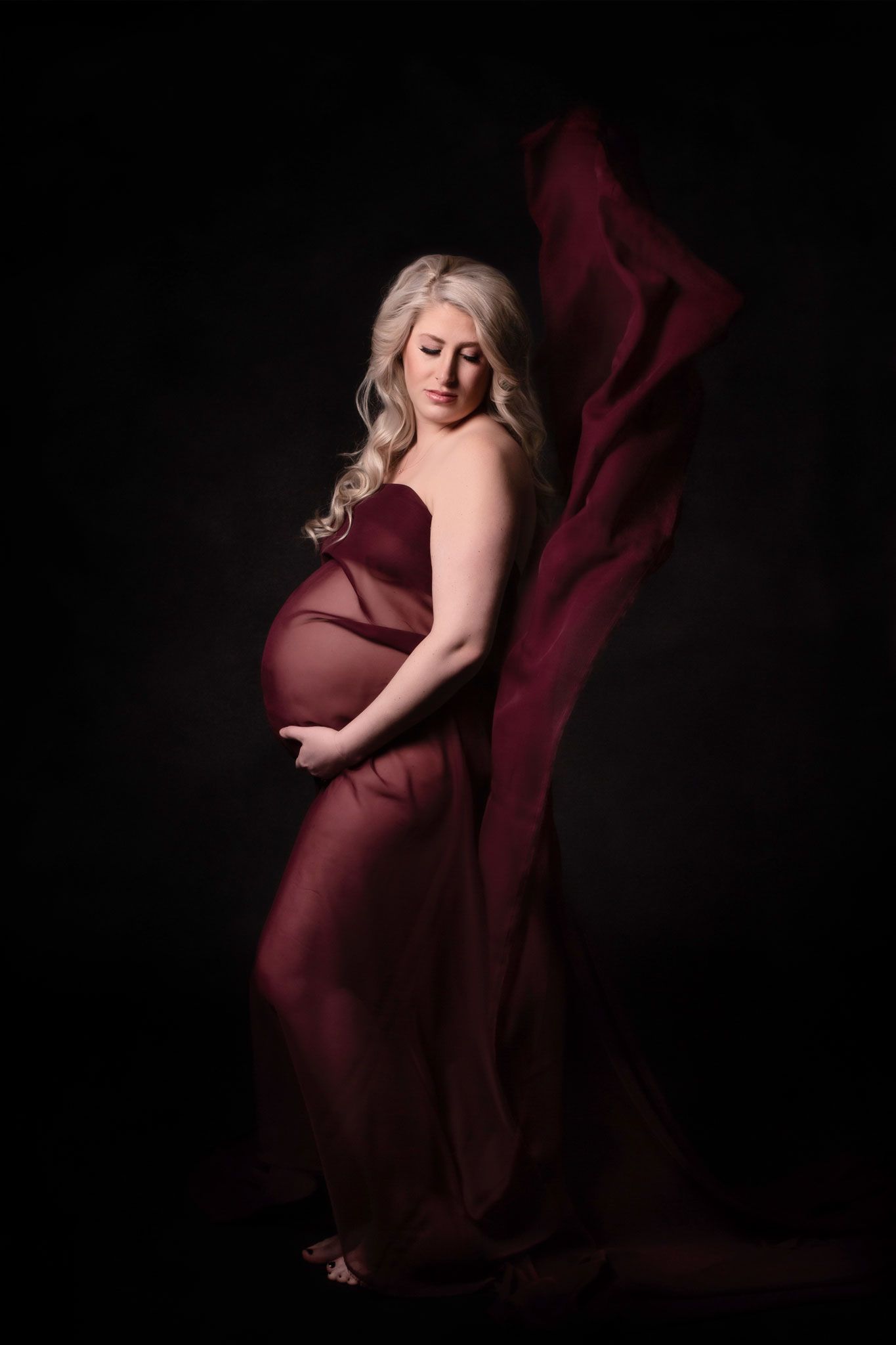 Artistic maternity photoshoot series in burgundy fabric against dark background showing pregnancy silhouette.