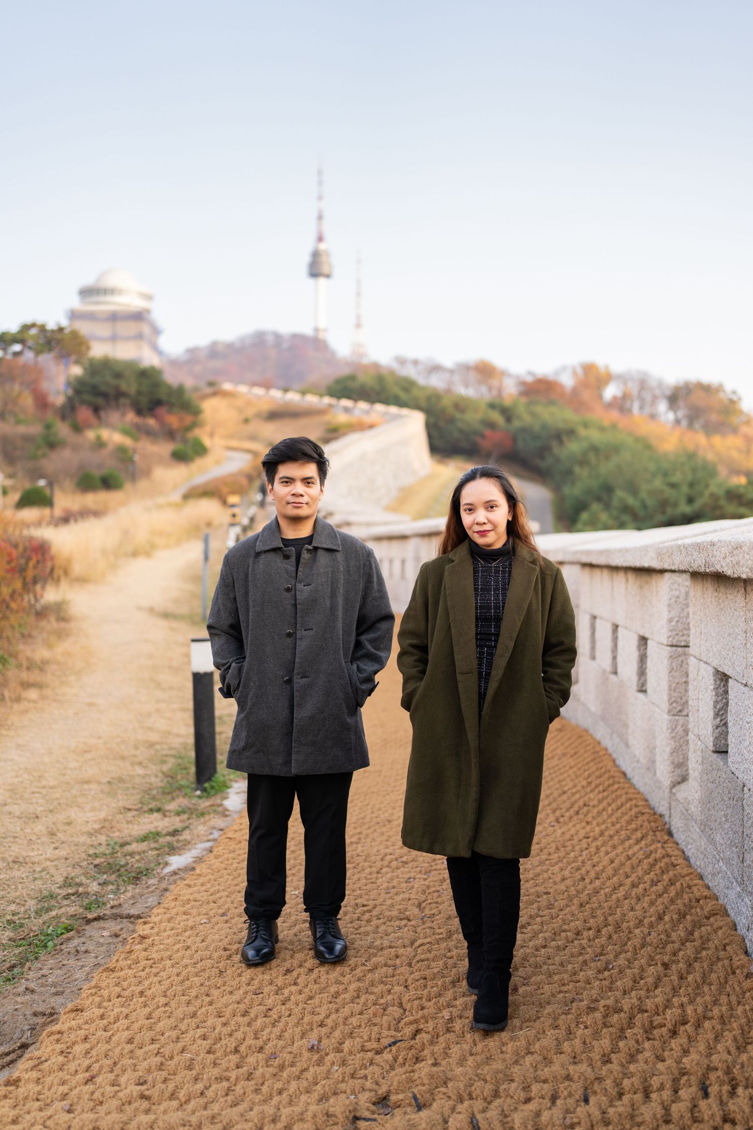 Two people in winter coats stand together on a winding path with Seoul Tower visible in the background.