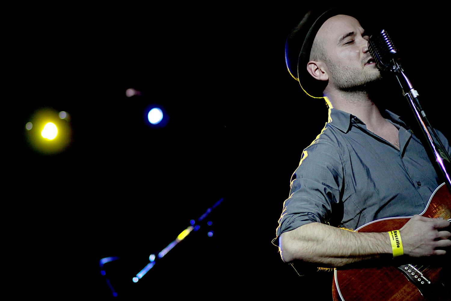 Male guitarist performing on stage with microphone wearing gray shirt and hat under concert spotlight.