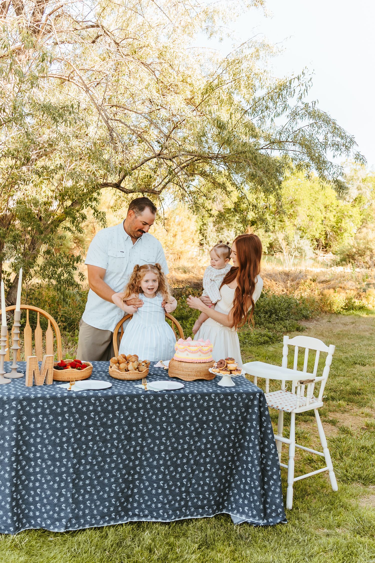 A family enjoys a festive outdoor picnic with desserts and treats laid out on a table with a blue polka dot tablecloth.