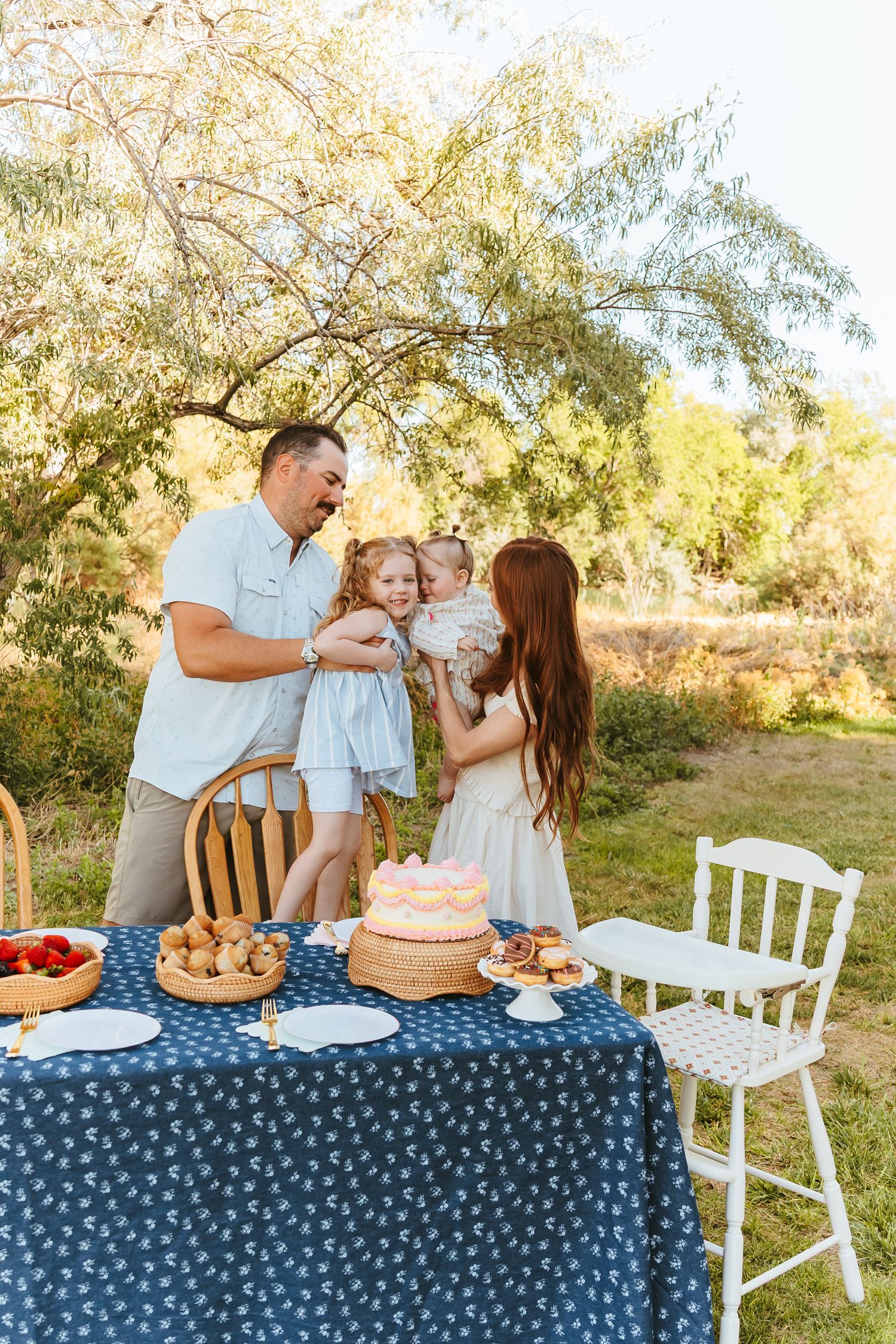Family enjoying an outdoor celebration with cake and treats on a blue polka dot tablecloth under a tree in golden sunlight.