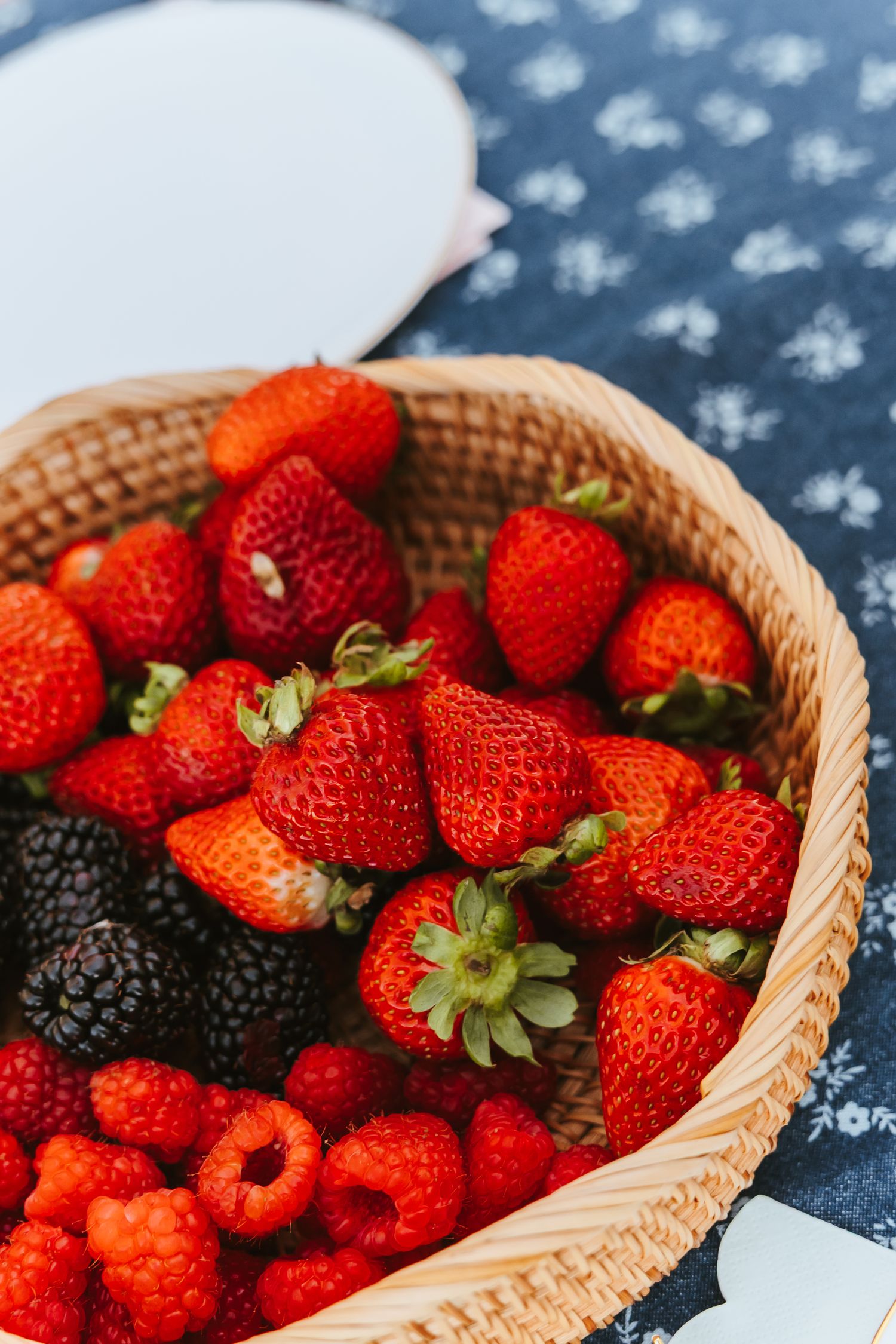 Fresh ripe strawberries and blackberries arranged in a woven basket against a dark blue textured background.