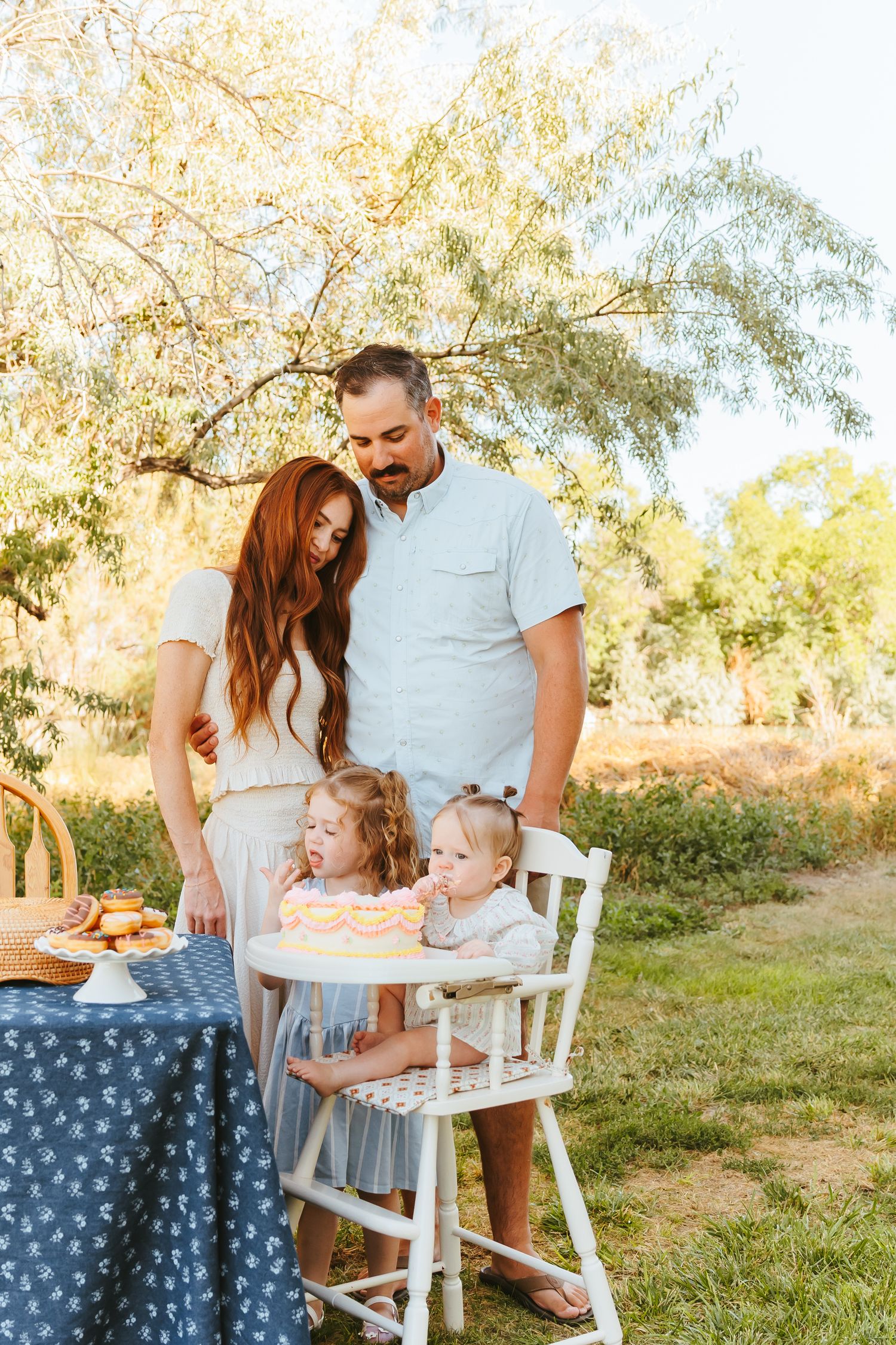 A family gathers around a white table for an outdoor celebration in a sunlit garden setting.