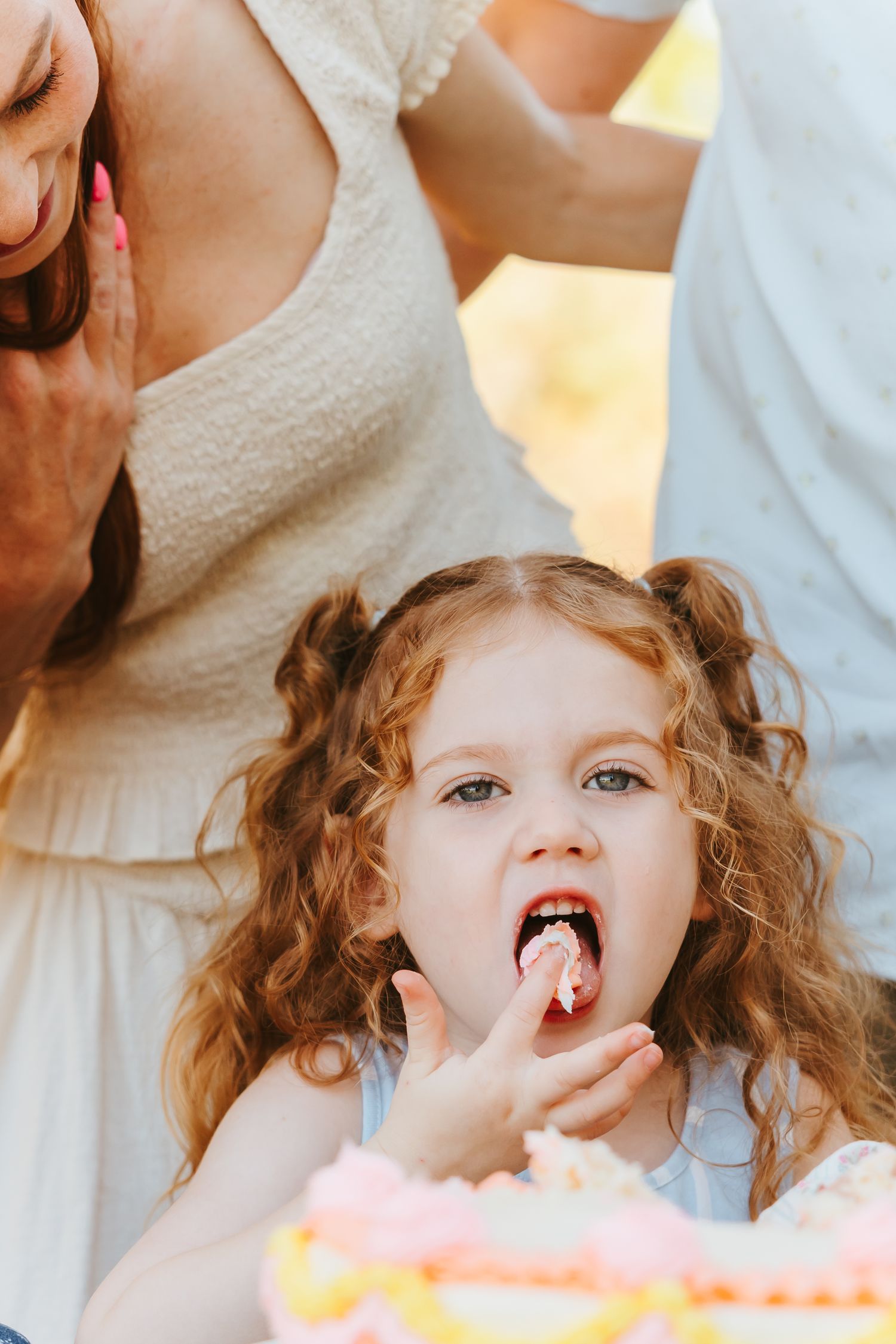 Young child with curly hair takes a bite of cake while wearing a white outfit.