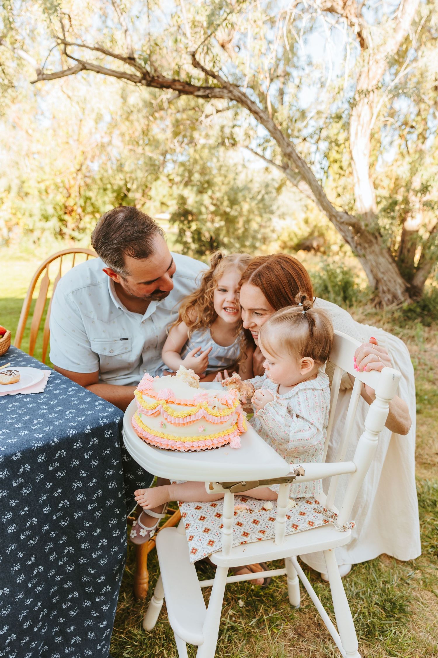 Family celebrates a birthday outdoors with cake at a table decorated with a blue tablecloth under a tree.