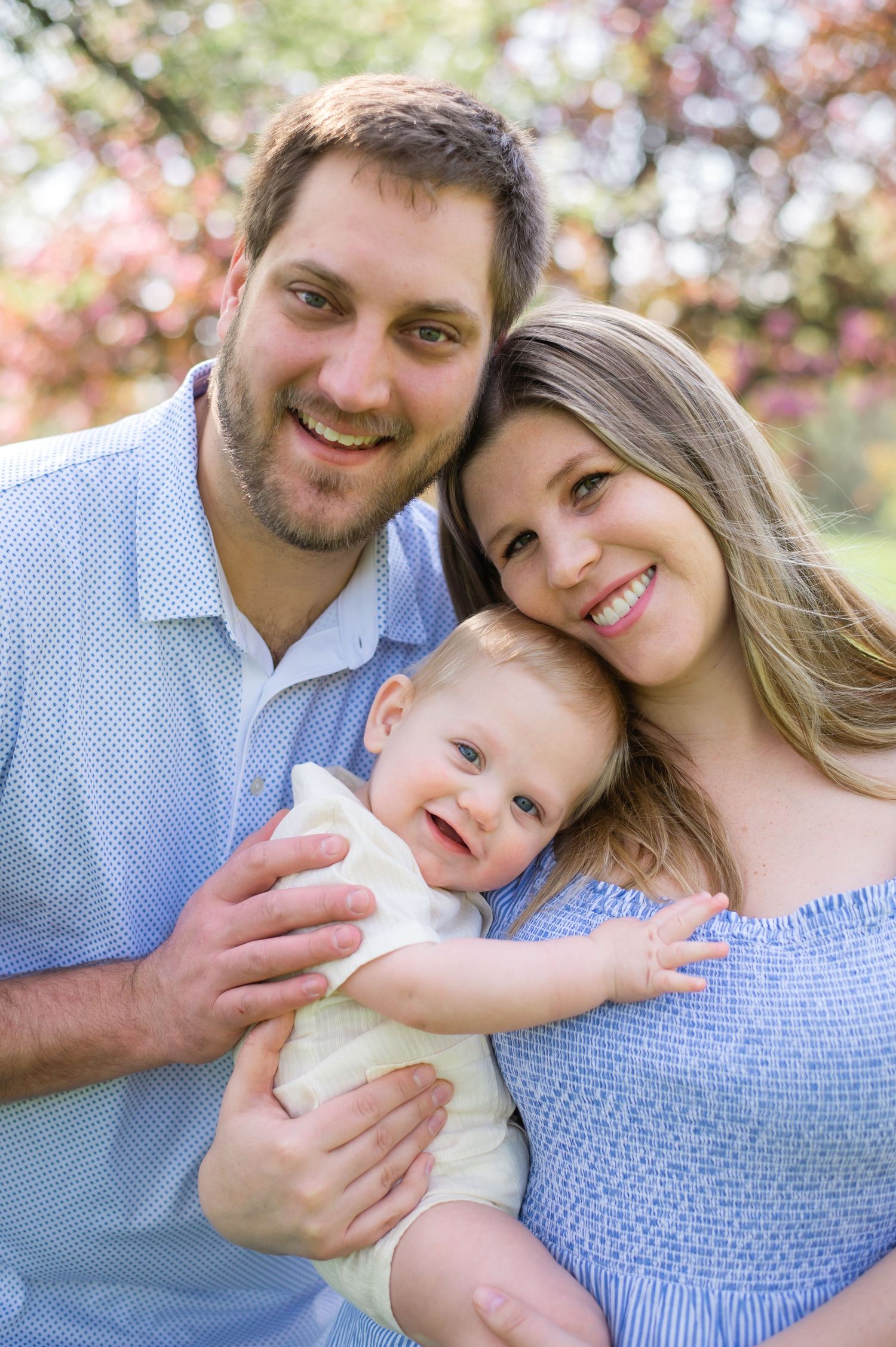 A happy family of three poses together outdoors on a sunny day with blurred foliage in the background.