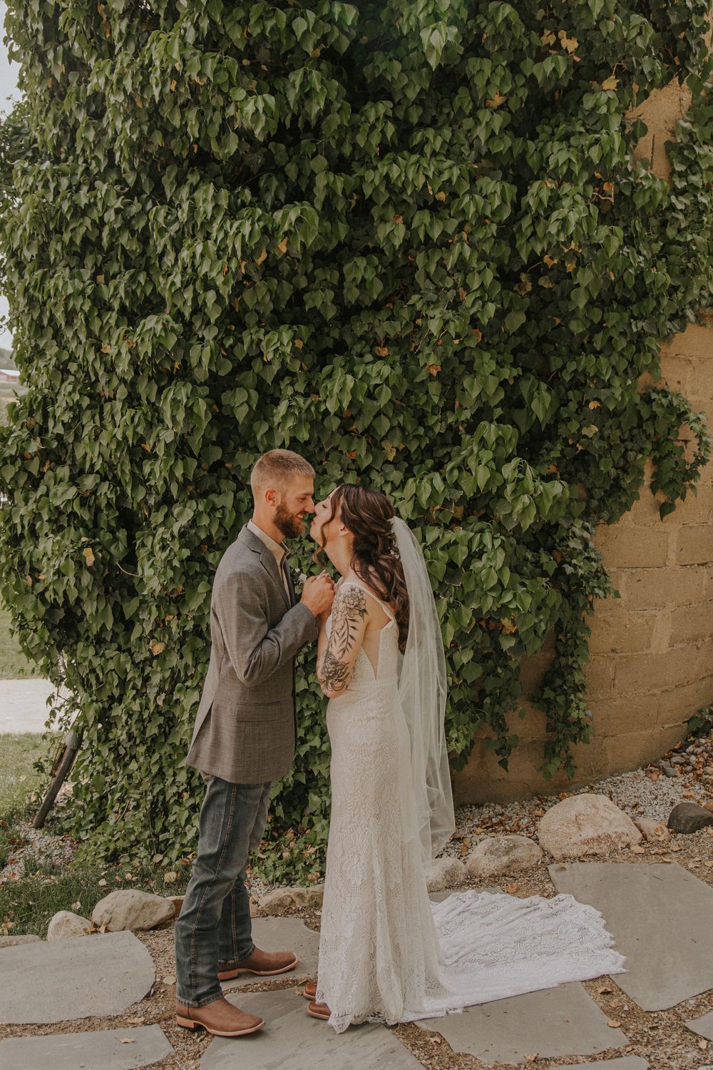 Bride and groom share a romantic moment in front of ivy-covered wall, bride in lace gown with veil.