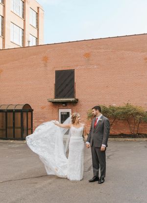 A bride's dress flows in the wind as she poses with her partner against a brick building.