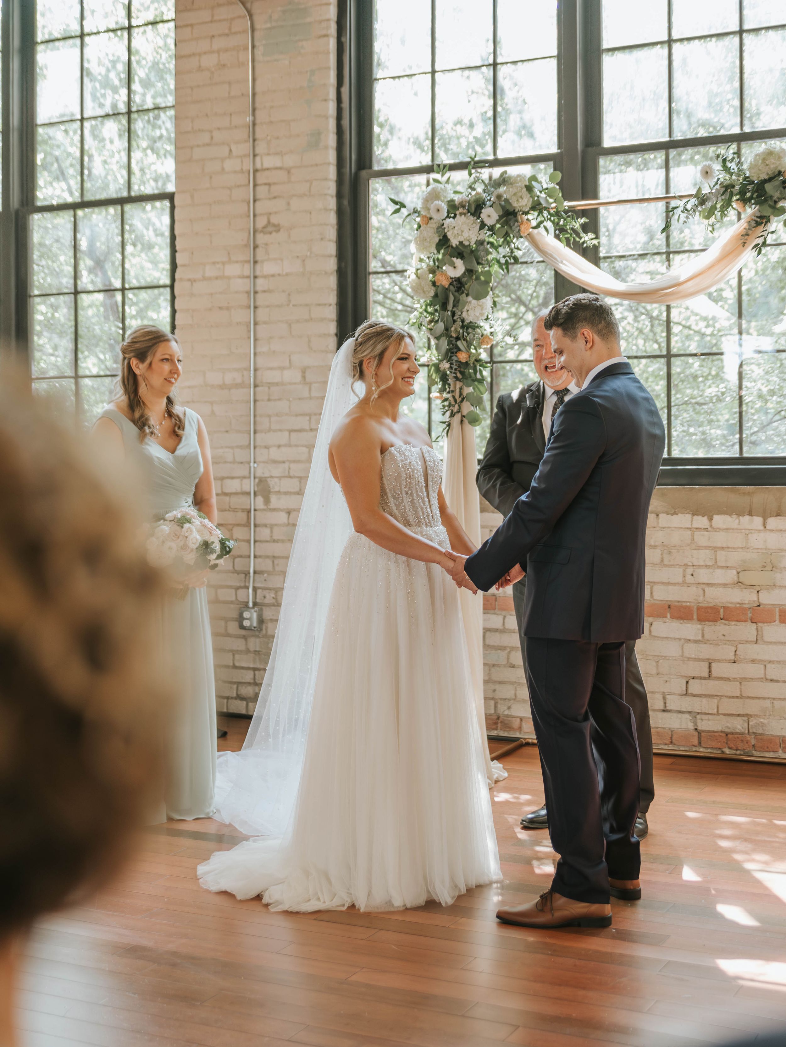 Bride and groom hold hands at indoor wedding ceremony with floral arch, brick walls, and bridesmaid in white dress.