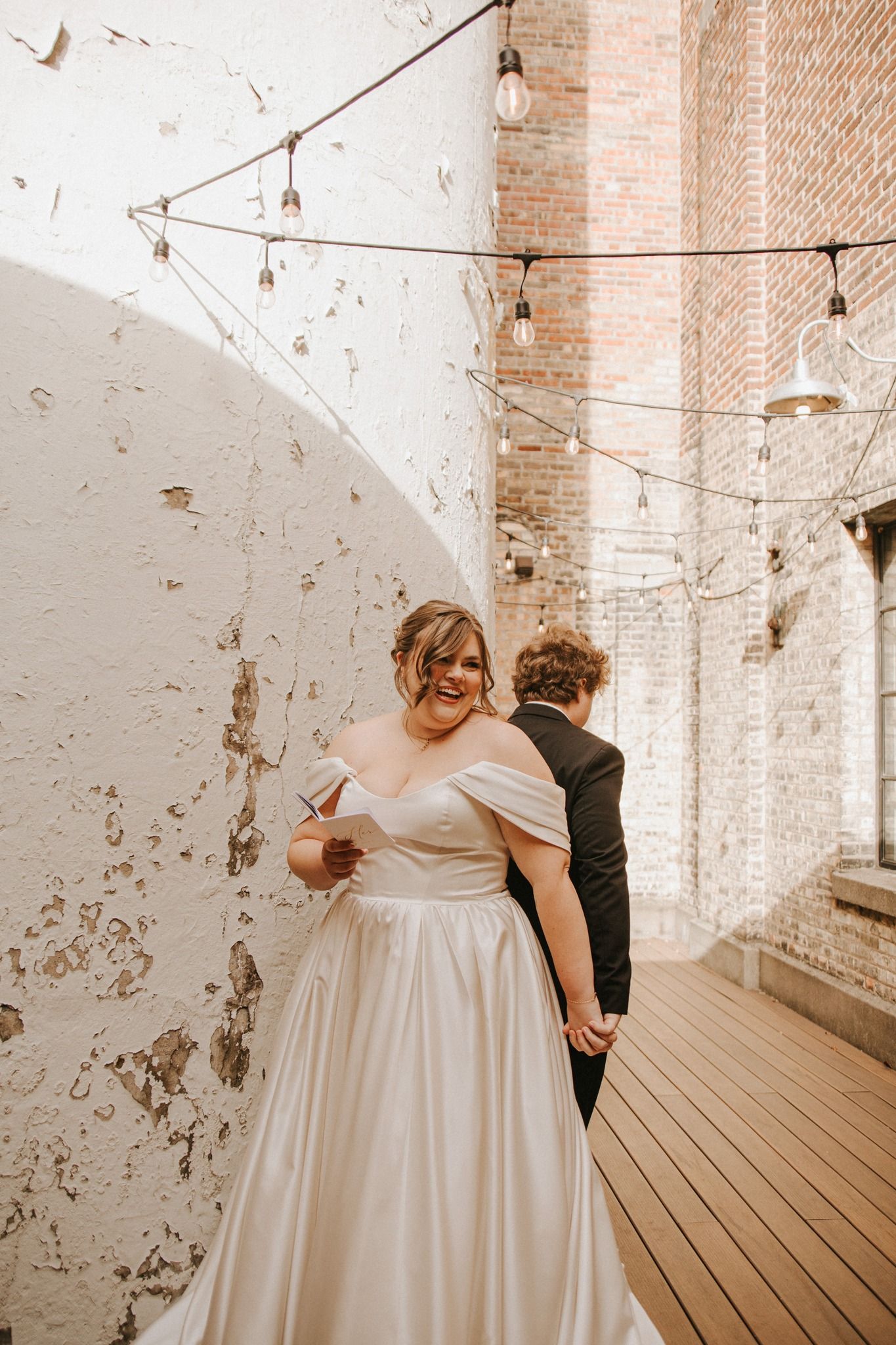 Bride in off-shoulder satin gown reads vows while groom stands behind her in industrial wedding venue with string lights.