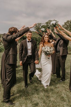 Smiling bride and groom walk hand-in-hand through an arch of raised hands formed by wedding party on green lawn.