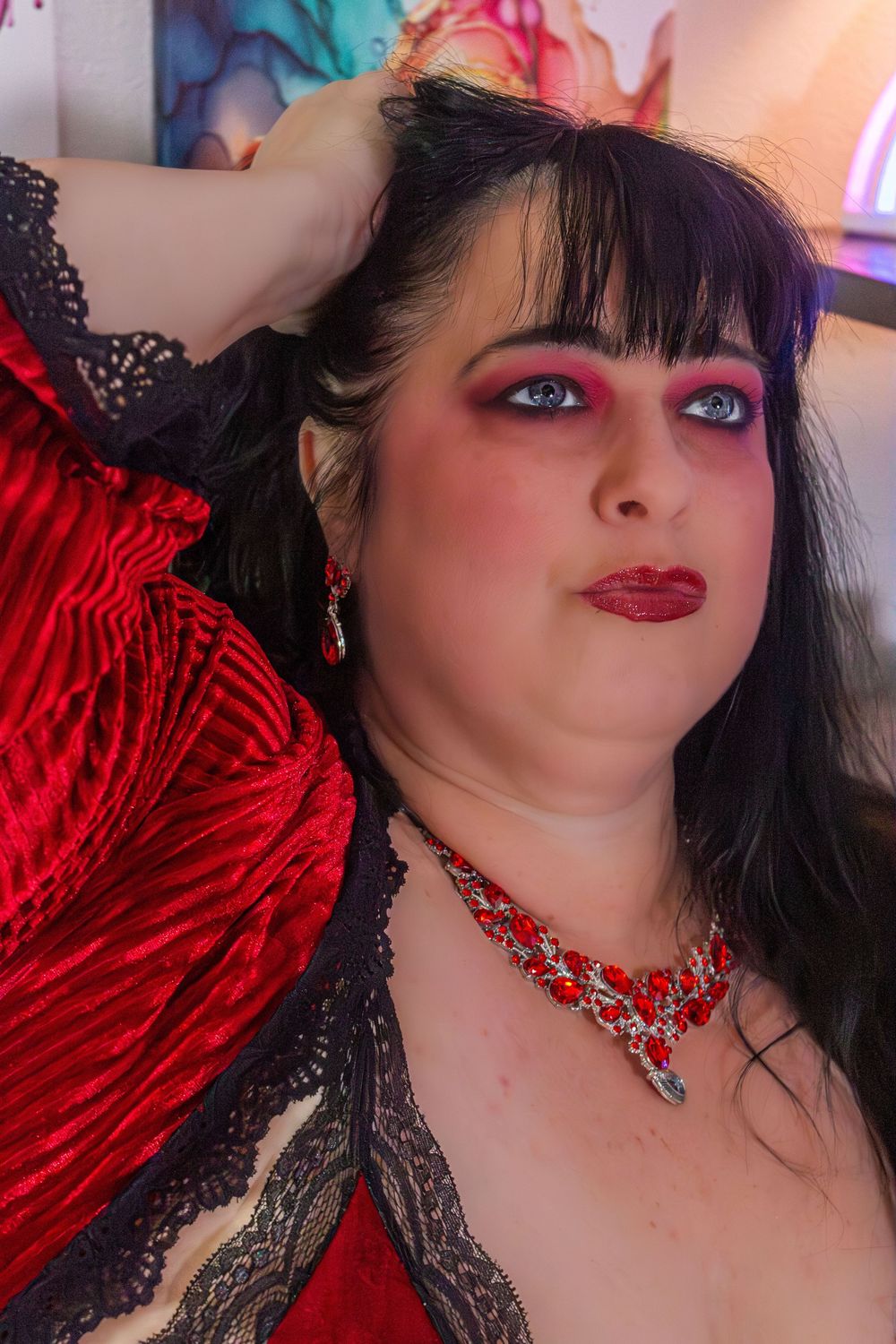 Woman in red velvet dress with black lace, red jewelry, dramatic makeup, posing confidently indoors.