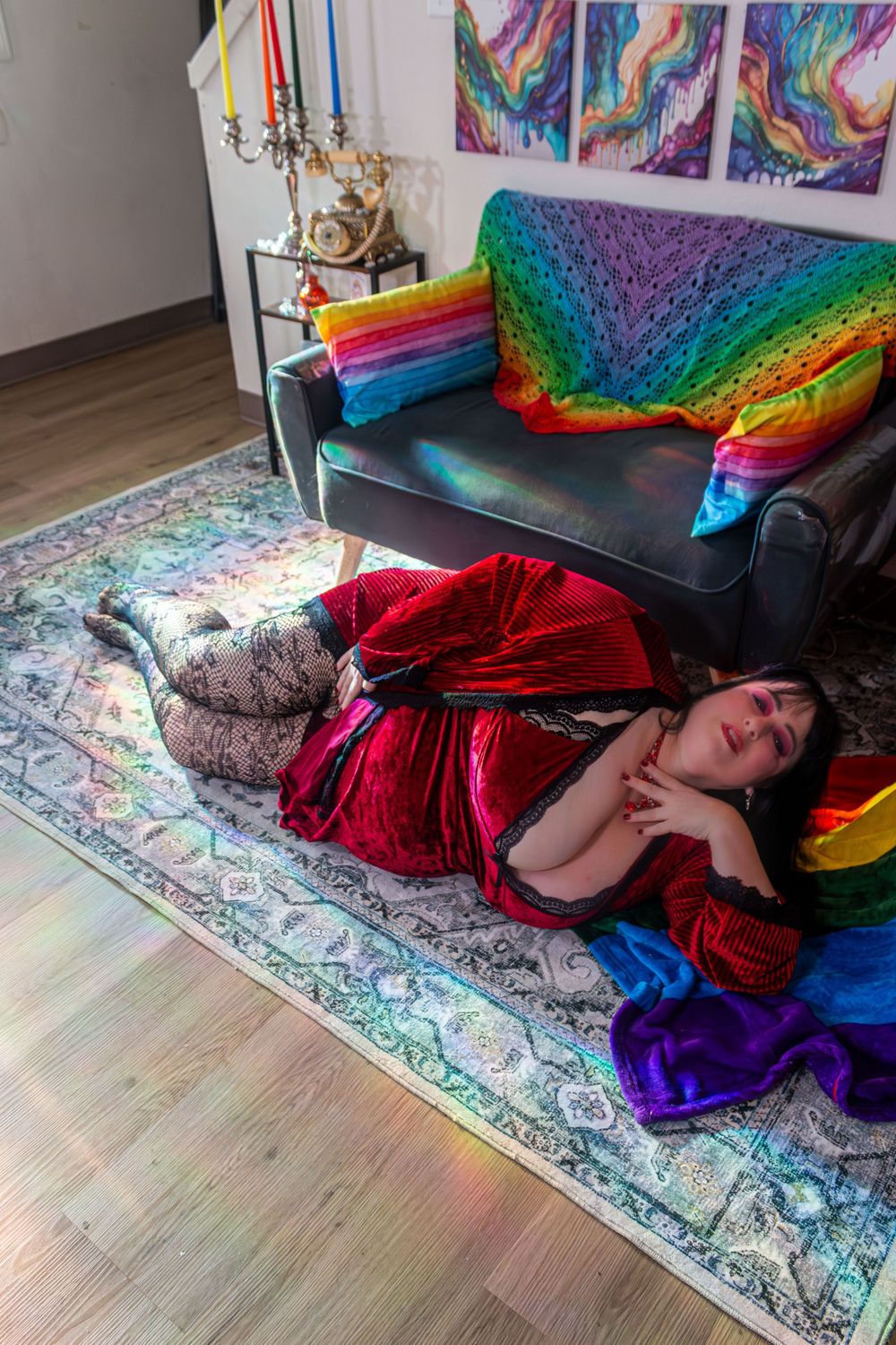 Woman in red velvet dress lying on colorful rug beside sofa with rainbow knit blanket in vibrant decorated room.