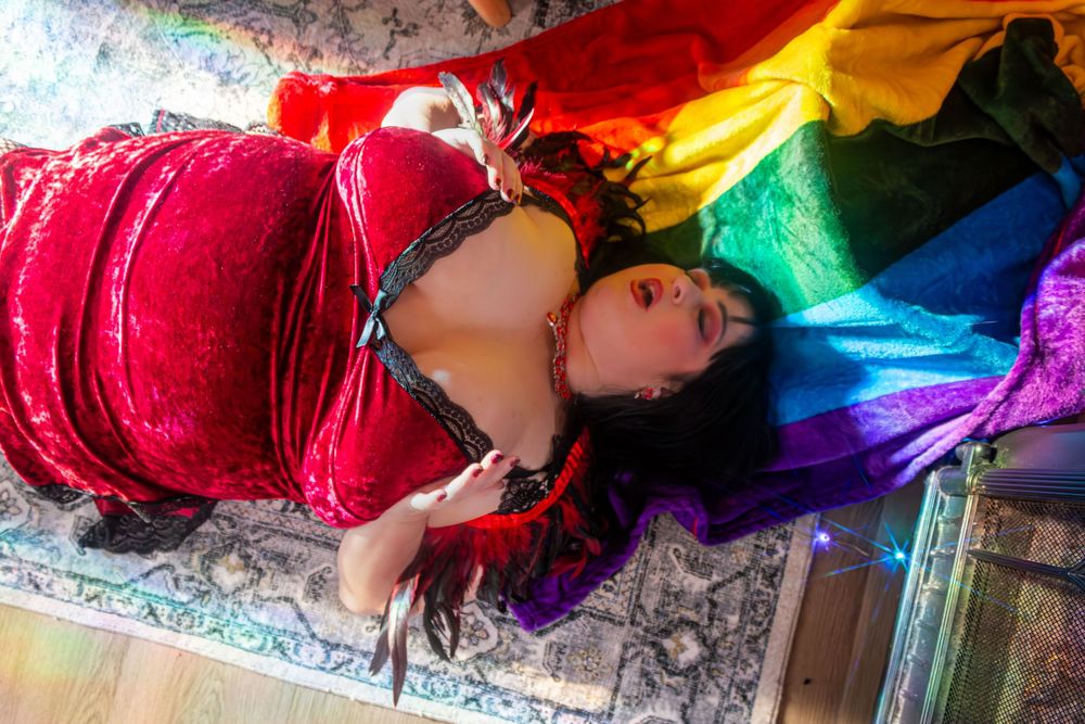 Woman in red velvet outfit lying on rainbow pride flag on patterned floor.