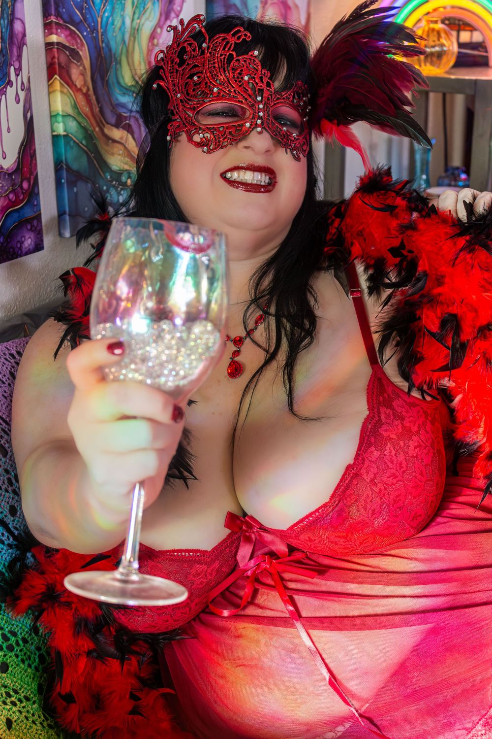 Woman in red lace lingerie and masquerade mask holding a wine glass, surrounded by colorful decor.