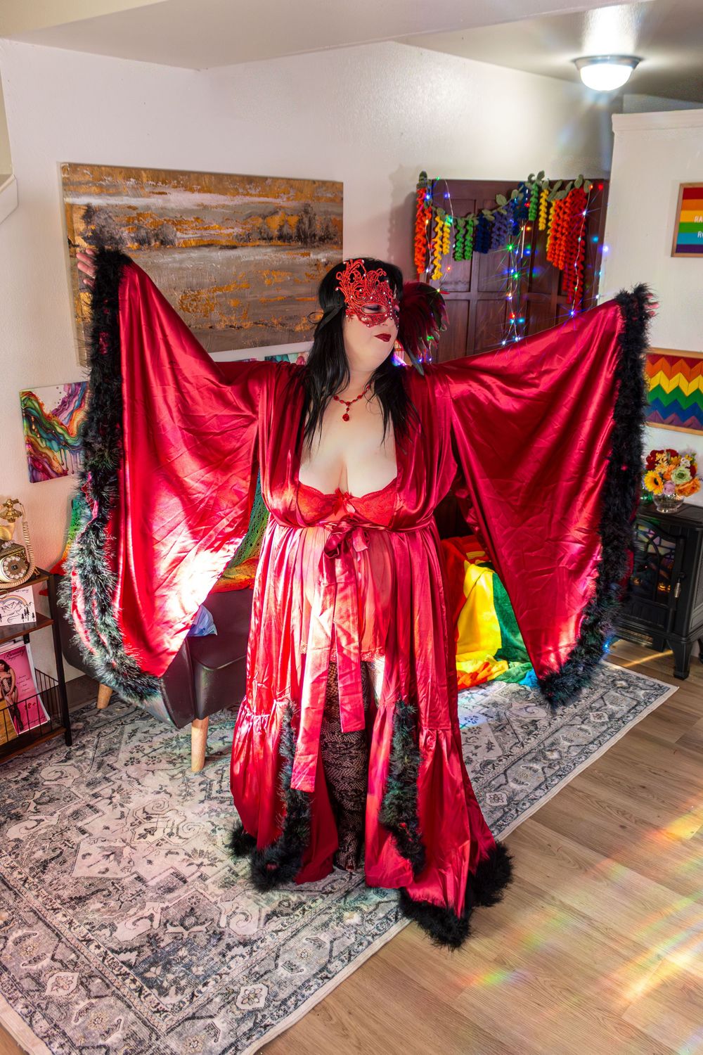 Woman in red satin robe with feather trim and masquerade mask poses in decorated room with rainbow pride flag.