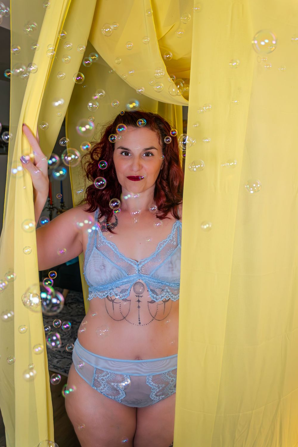Woman in blue lace lingerie stands between yellow curtains surrounded by floating soap bubbles.