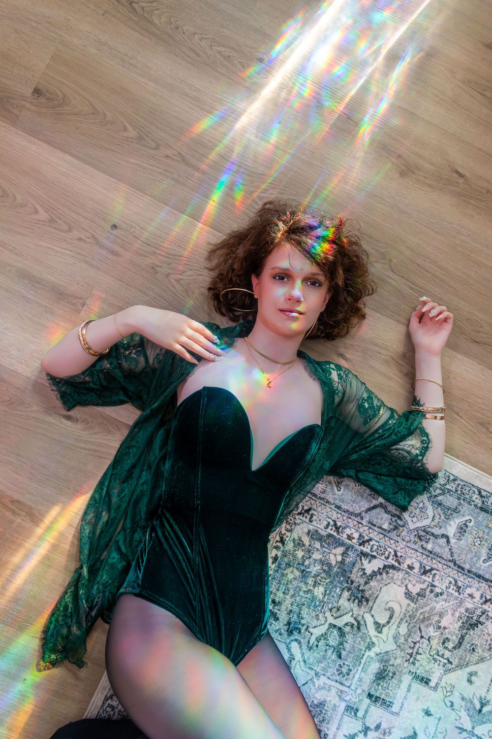 Woman in green velvet bodysuit lying on hardwood floor with rainbow light prism reflections.