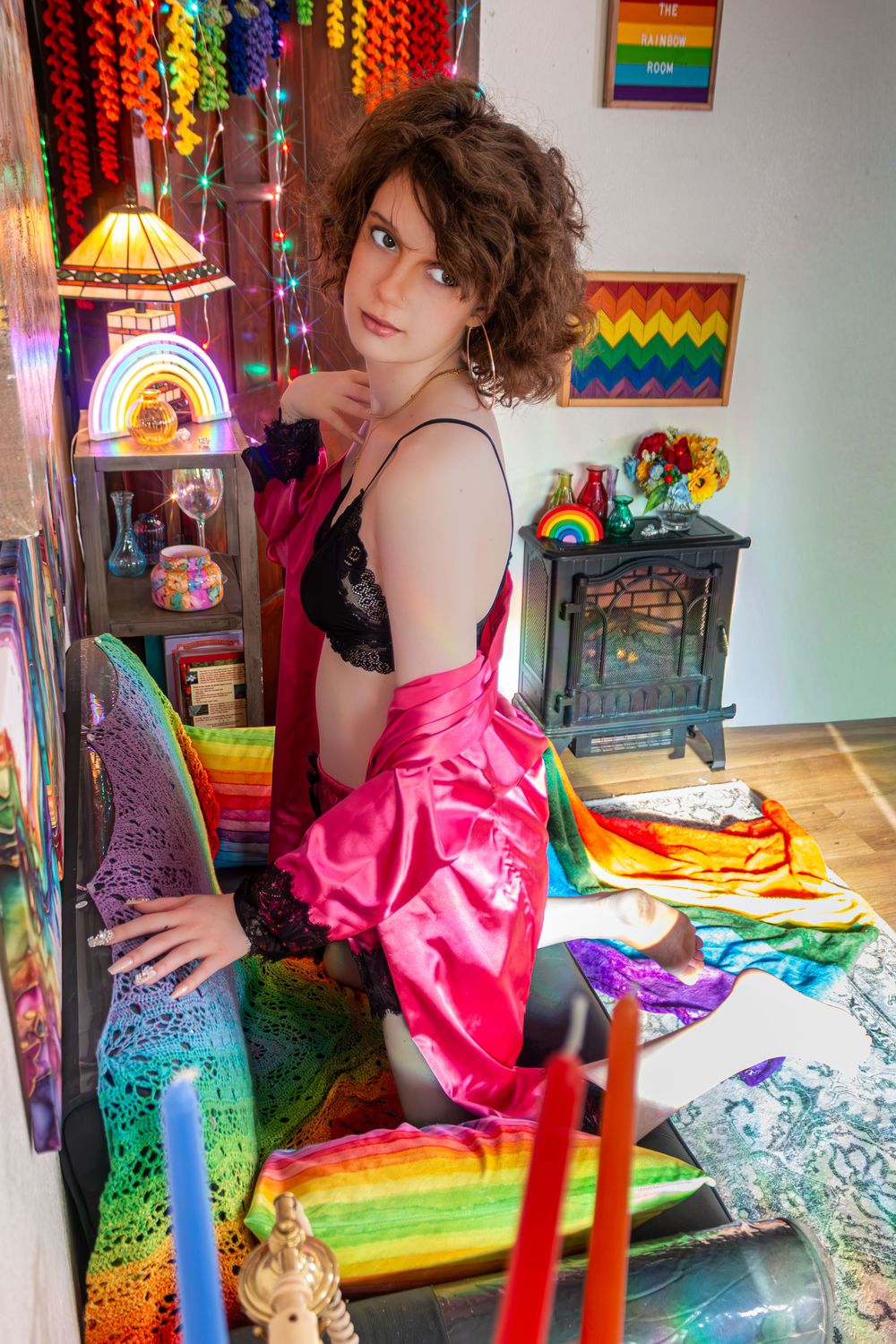 Person in pink satin skirt and black top poses in colorful pride-themed room with rainbow decorations.