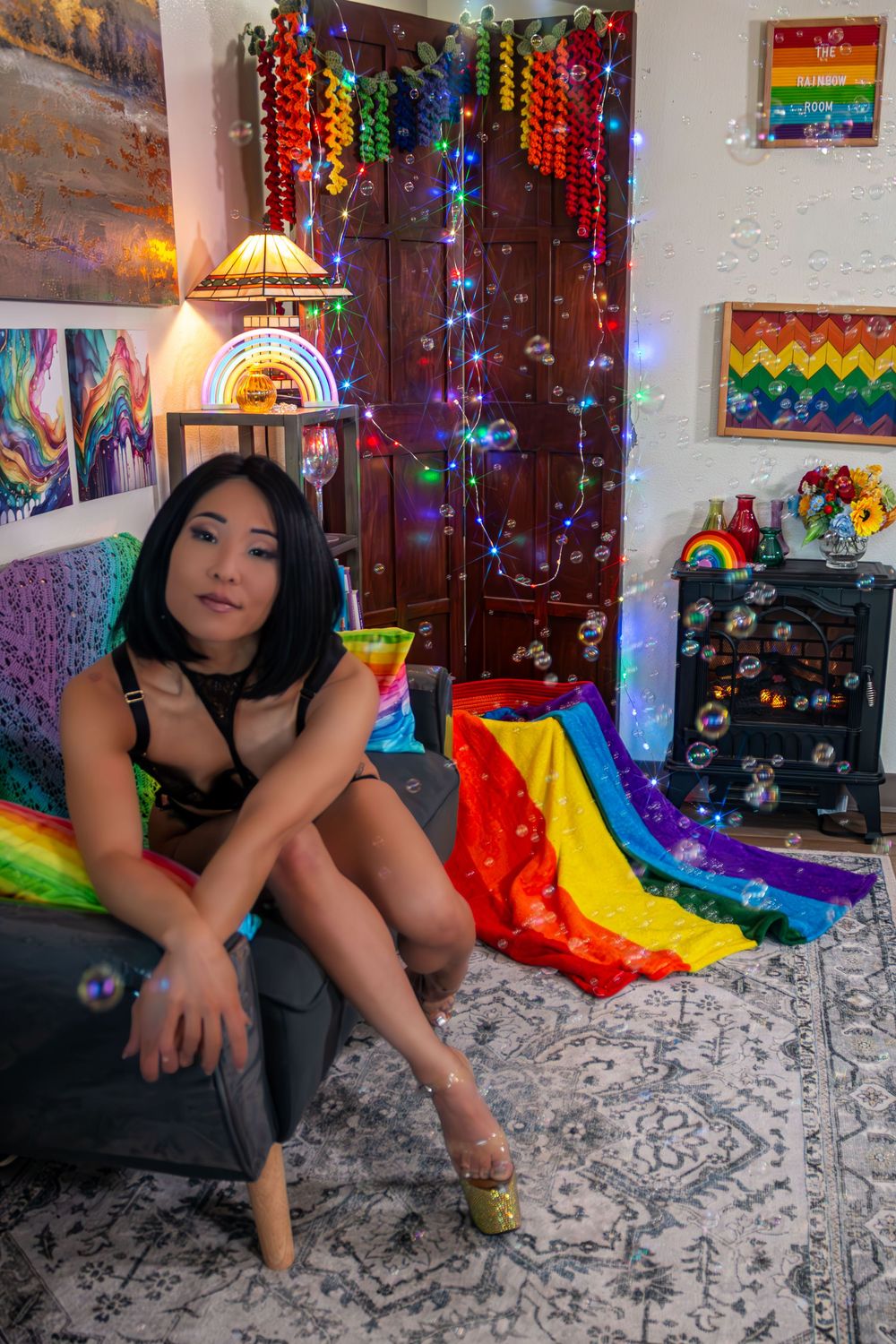 Woman sitting in a colorful Pride-decorated room with rainbow flags, fairy lights, and vibrant artwork on the walls.