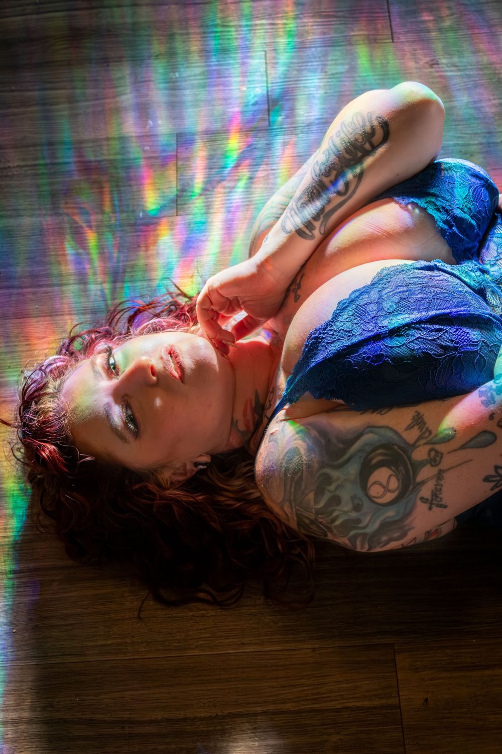 Artistic portrait with rainbow light effects showing person with tattoos and red hair in blue lingerie lying on wooden floor.