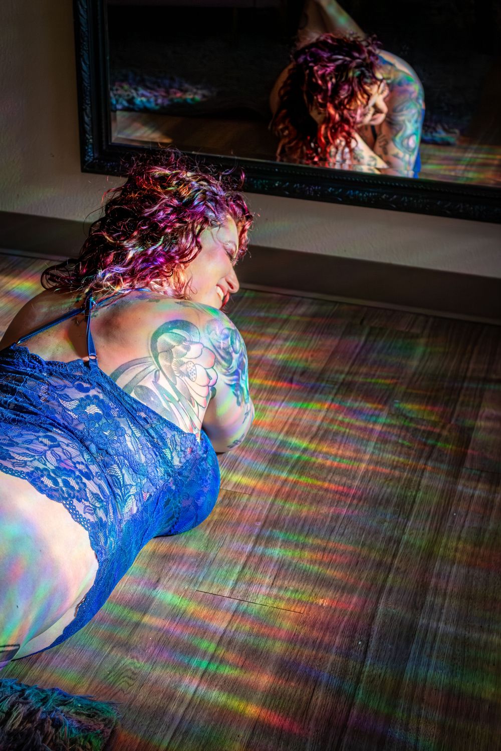 A person in a blue lace bodysuit lies on a hardwood floor as rainbow light patterns reflect across the scene.