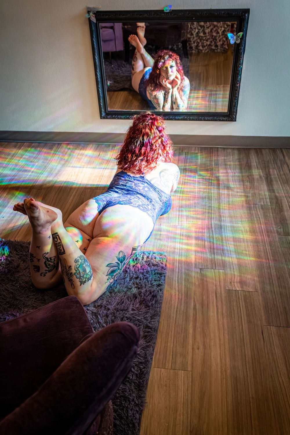 Mirror reflection shot of person with red hair and tattoos in blue lingerie on hardwood floor with prismatic lighting effects.