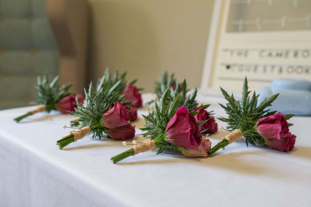 Wedding boutonnieres with deep red roses and greenery arranged on a white surface.
