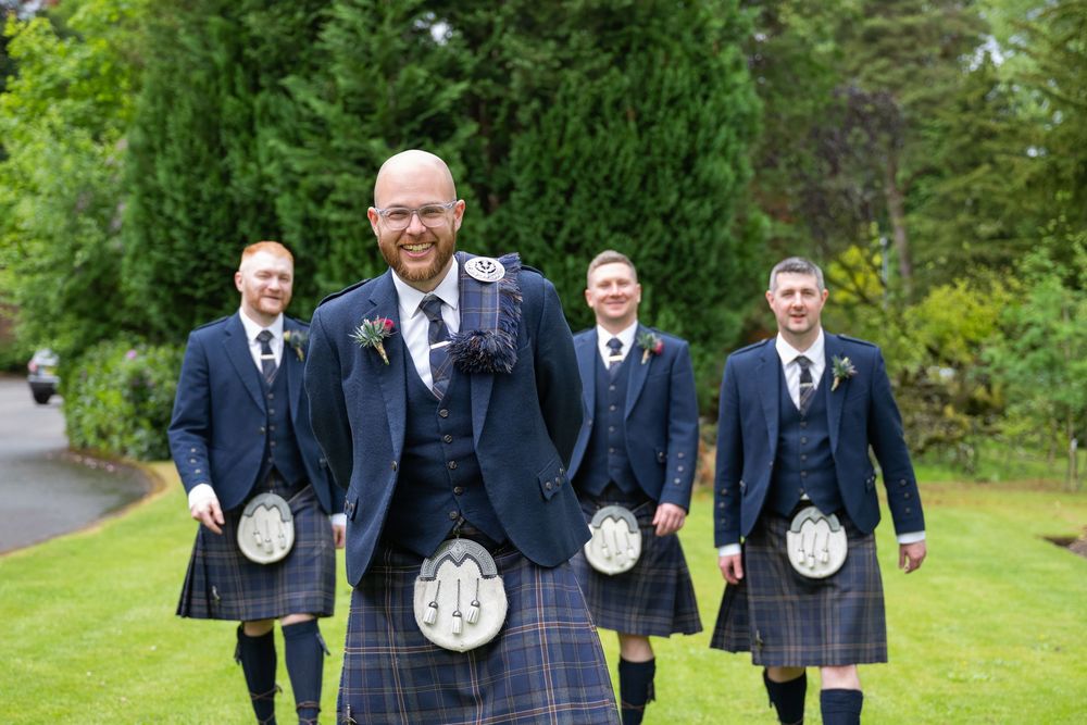 Wedding party in traditional Scottish kilts and navy jackets pose outdoors.