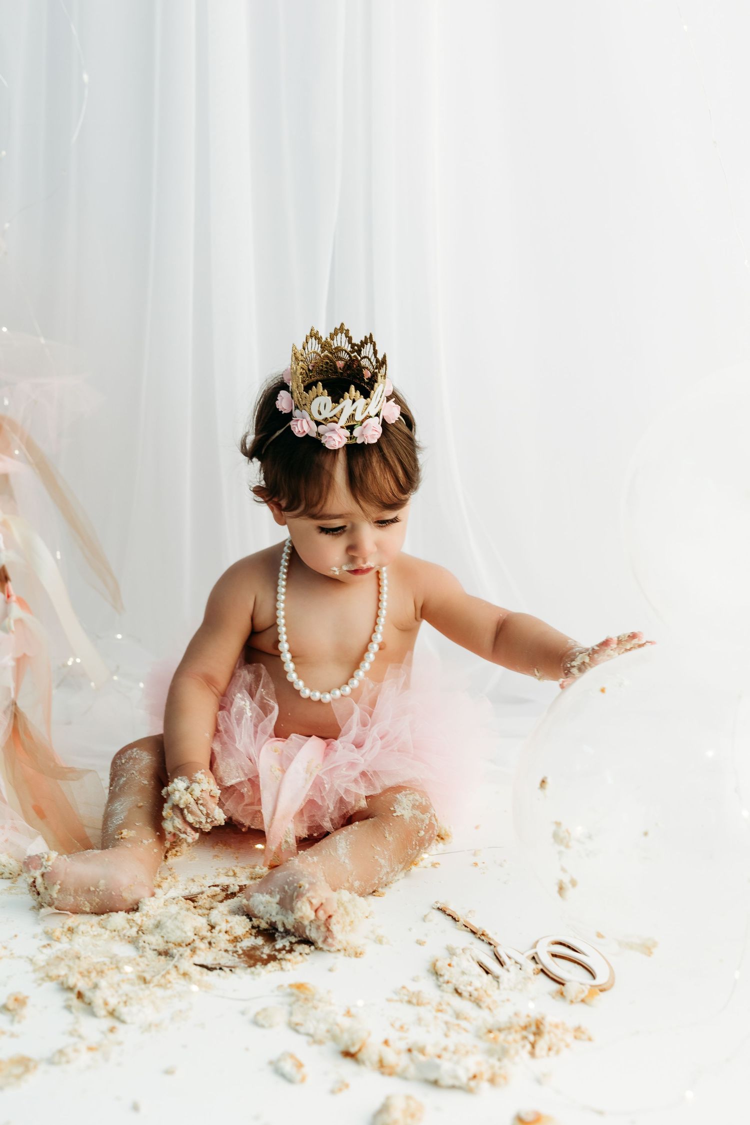 A small child in a white lace dress sits on the floor playing with cake and frosting.