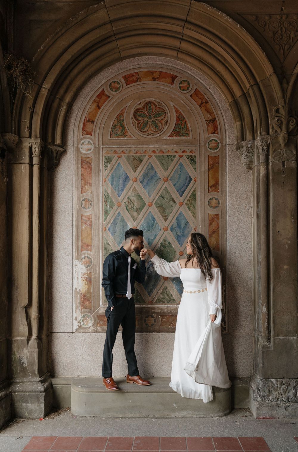 Couple sharing a romantic moment under an ornate stone arch with colorful frescoes at Bethesda Terrace, Central Park.