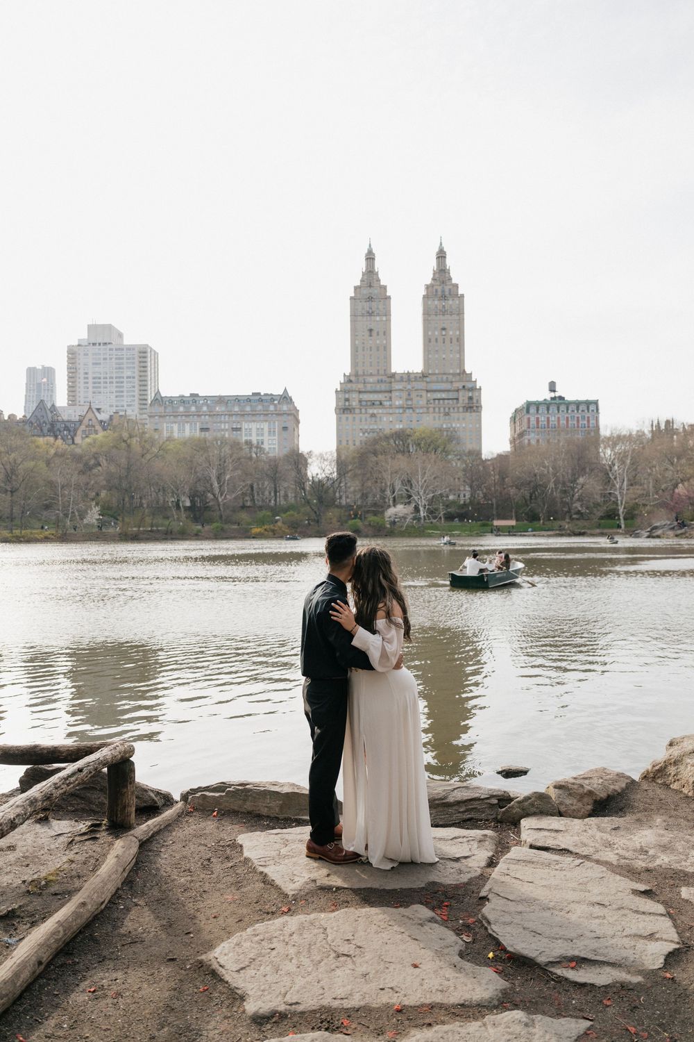 Bride and groom embracing by the lake in Central Park with the San Remo building skyline in the background.