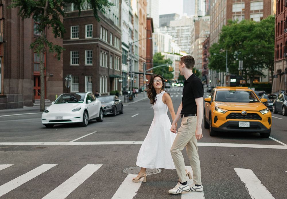 Couple holding hands crossing a NYC street crosswalk, yellow taxi and Tesla visible, woman in white dress.