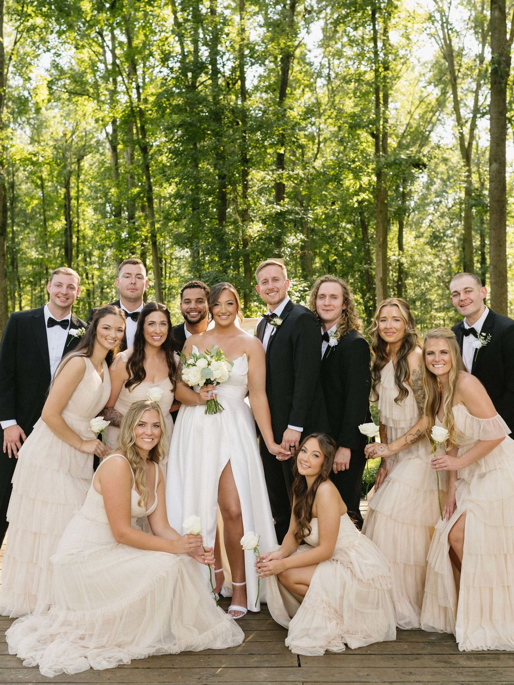 Wedding party in white dresses and black tuxedos posing together on wooden deck.