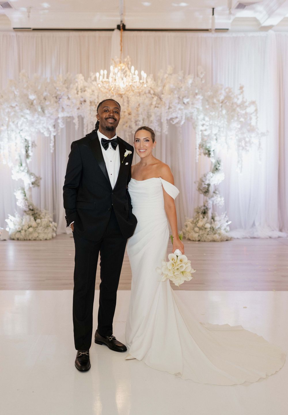 Bride and groom pose elegantly in white gown and black tuxedo before a stunning white floral backdrop.