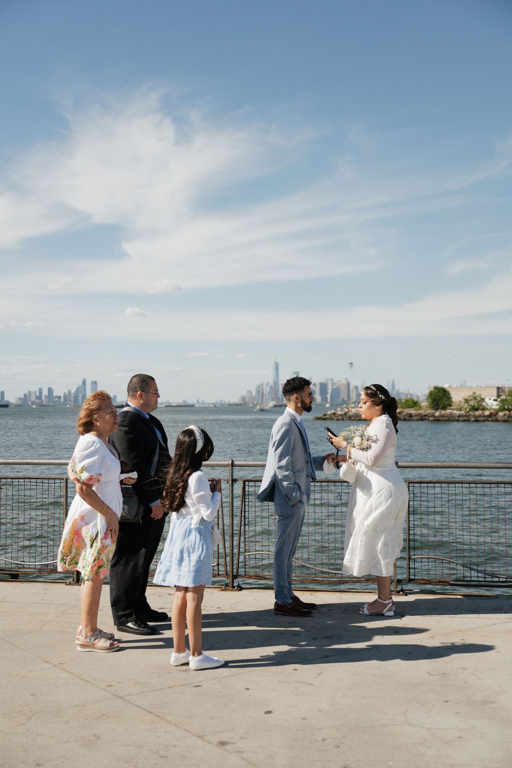 Small wedding ceremony on NYC waterfront with Manhattan skyline, couple exchanging vows with family on sunny day.