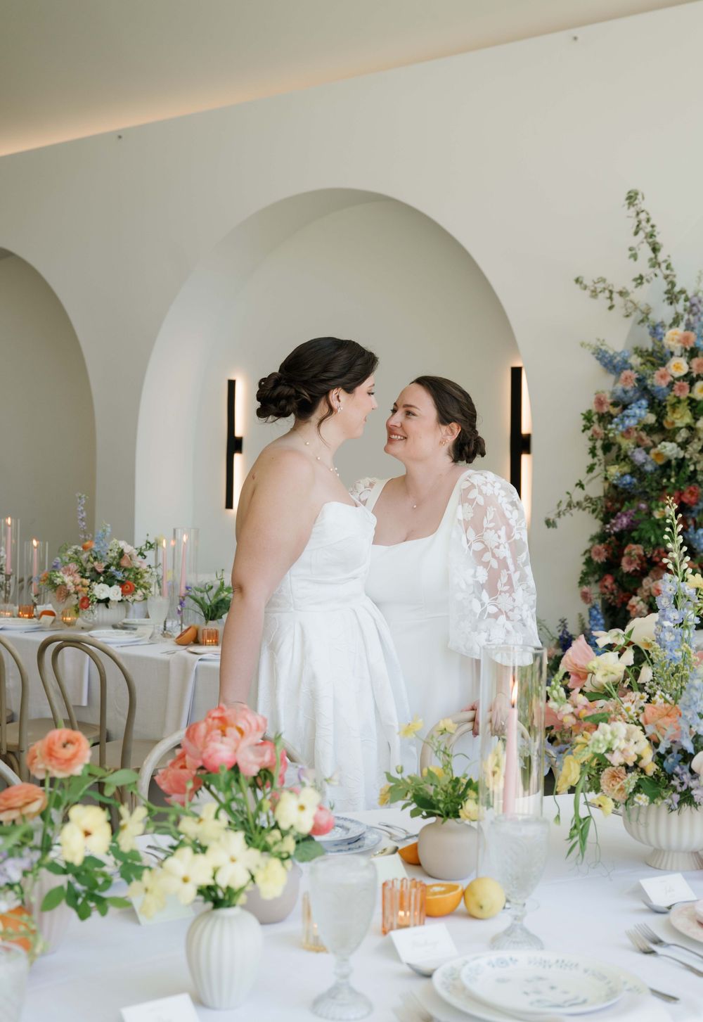 Two brides in white gowns share a tender moment at a floral-adorned wedding reception table with colorful blooms.
