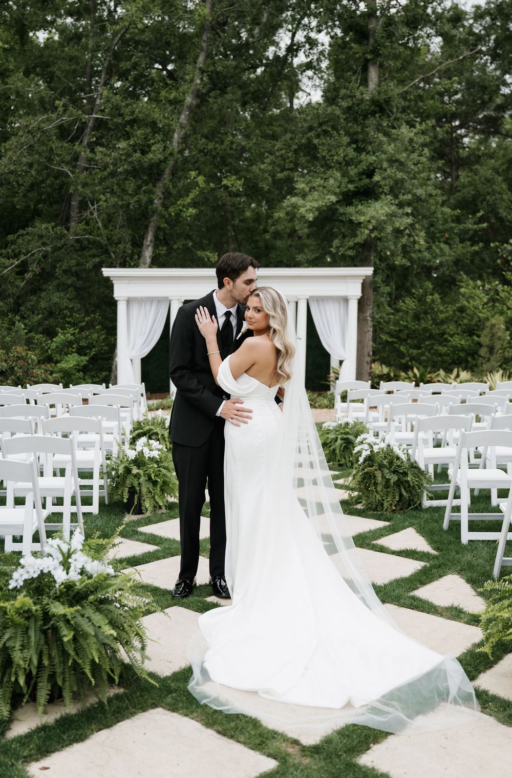 Bride and groom pose at outdoor wedding ceremony with white chairs, floral greenery, and elegant white arbor backdrop.