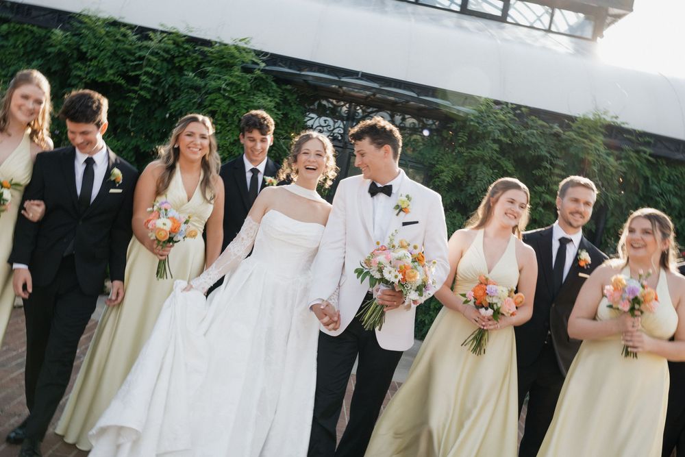Wedding party in black tuxedos and gold bridesmaid dresses walks with bride and groom holding bouquets outdoors.